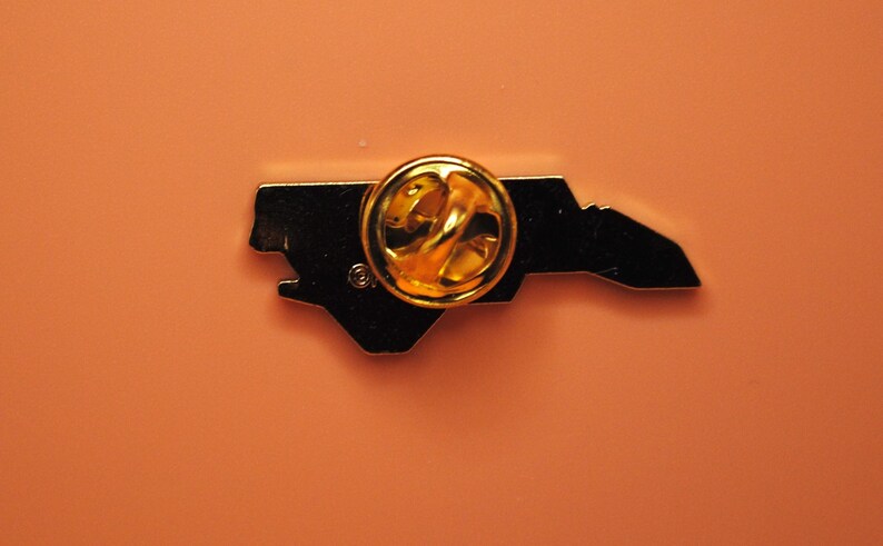 May include: A black enamel pin in the shape of the state of North Carolina, with a gold-coloured pin back. The pin is set against an orange background.