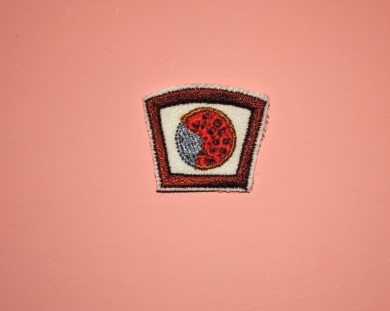 May include: A red and white embroidered patch with a slice of pizza inside a brown frame.