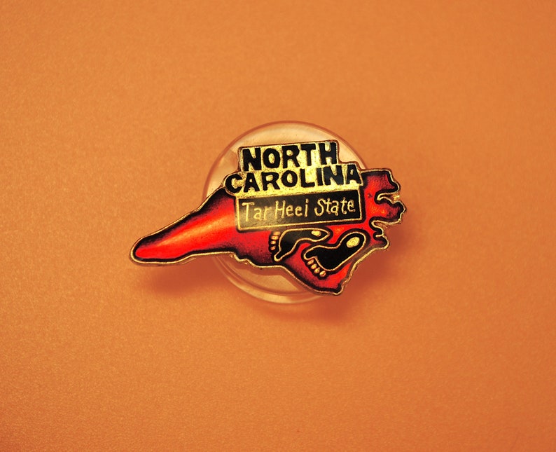 May include: A red and gold enamel pin shaped like the state of North Carolina with the words "North Carolina" and "Tar Heel State" in black lettering.