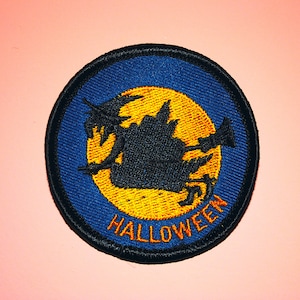 May include: Embroidered patch with a Halloween theme. The patch features a black silhouette of a witch on a broomstick against a yellow moon, set within a blue circle. The word "HALLOWEEN" is stitched in orange.