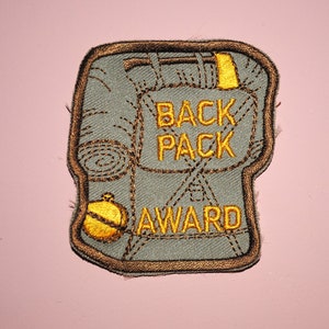 May include: A gray embroidered patch with a brown outline. The patch depicts a backpack with the text "BACK PACK AWARD" in yellow thread.
