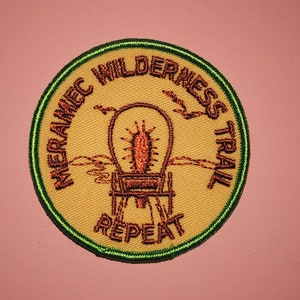 May include: A round, yellow embroidered patch with a green border. The patch features a wagon with a red and brown design and the text "Meramec Wilderness Trail Repeat".