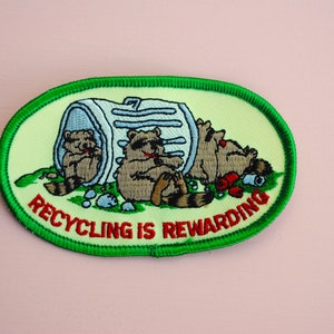 May include: Green and white embroidered patch with three raccoons in a trash can. The text "Recycling is Rewarding" is written in red.