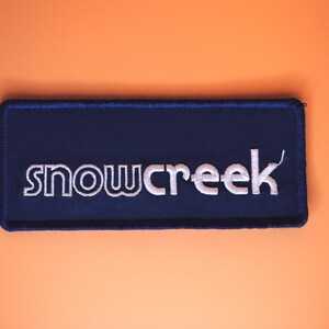 May include: A navy blue rectangular patch with the word "snowcreek" embroidered in white. The patch has a dark blue border and is set against an orange background.