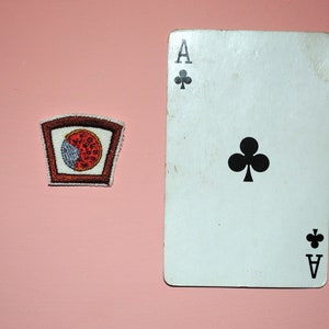 May include: A white playing card with a black ace of clubs symbol. The card is on a pink surface next to a brown and red embroidered patch with a pizza slice design.
