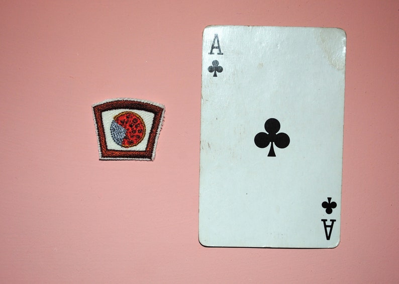 May include: A white playing card with a black ace of clubs symbol. The card is on a pink surface next to a brown and red embroidered patch with a pizza slice design.