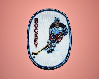 May include: A white oval patch with a blue border. The patch features a hockey player in a blue and white uniform skating with a stick and puck. The word "HOCKEY" is embroidered in red on the left side of the patch.