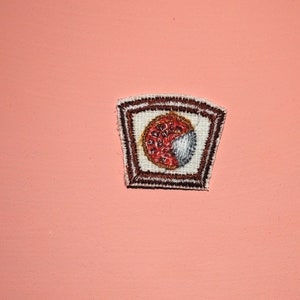 May include: A brown and white embroidered patch with a red and white circle design in the center.