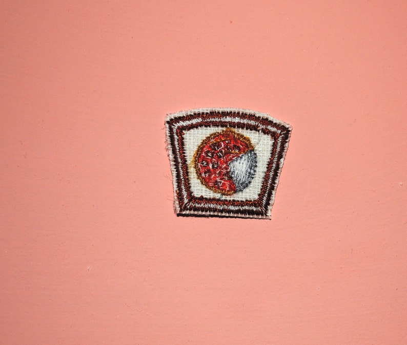May include: A brown and white embroidered patch with a red and white circle design in the center.