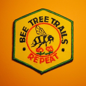 May include: A yellow hexagonal patch with a green border. The patch features a bee with black and yellow stripes flying over two red flowers. The text "BEE TREE TRAILS" is written in a black circular border with an orange outline. The text "REPEAT" is written in a red circular border with an orange outline.