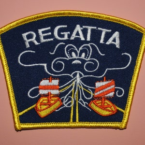 May include: A blue and gold embroidered patch with the word "REGATTA" in white lettering. The patch features a stylized image of two sailboats with red and white striped sails, sailing under a white cloud.