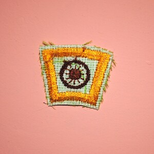 May include: Embroidered patch with a square design featuring a brown circular emblem with radiating lines, set within a gold border. The patch has a light green background and a frayed edge. The patch is set against a pink background.