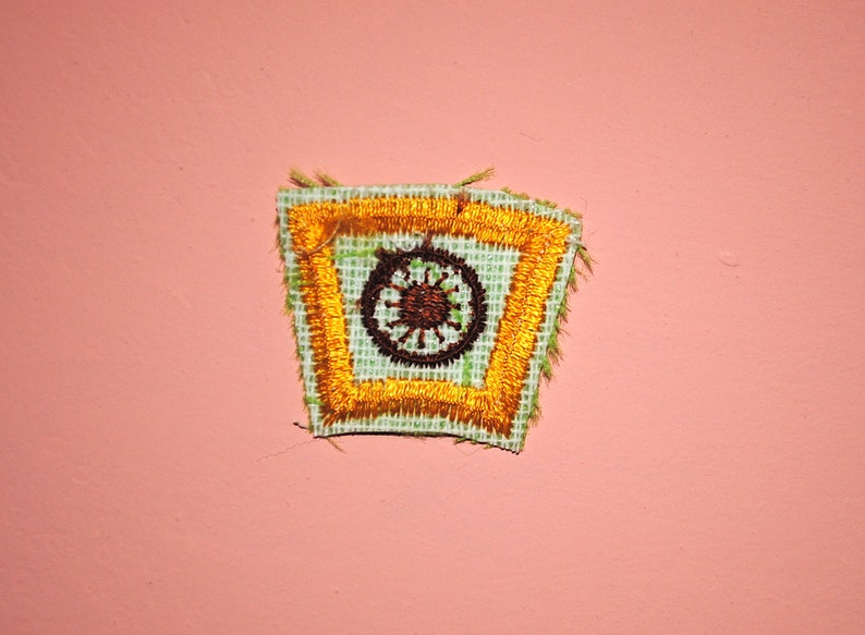 May include: Embroidered patch with a square design featuring a brown circular emblem with radiating lines, set within a gold border. The patch has a light green background and a frayed edge. The patch is set against a pink background.