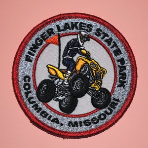 May include: A red, white, and gray embroidered patch with the text "Finger Lakes State Park Columbia, Missouri". The patch features a yellow ATV with a rider in a black helmet and blue and black clothing.