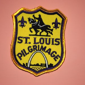 May include: A yellow and purple embroidered patch with a shield shape. The patch features a knight on horseback, two fleur-de-lis, and the text "St. Louis Pilgrimage".