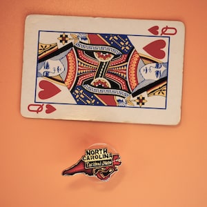 May include: A playing card, the Queen of Hearts, is lying on a peach-colored surface. A small, red, white, and blue enamel pin shaped like the state of North Carolina is below the card. The pin says "North Carolina Tar Heel State" in white letters.