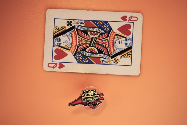 May include: A playing card, the Queen of Hearts, is lying on a peach-colored surface. A small, red, white, and blue enamel pin shaped like the state of North Carolina is below the card. The pin says "North Carolina Tar Heel State" in white letters.