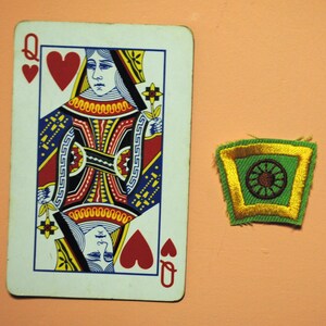 May include: A Queen of Hearts playing card and a green and gold embroidered patch with a brown circular design. The card features a detailed illustration of a queen in red, black, yellow, and blue. The patch has a gold border.