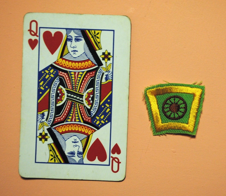May include: A Queen of Hearts playing card and a green and gold embroidered patch with a brown circular design. The card features a detailed illustration of a queen in red, black, yellow, and blue. The patch has a gold border.