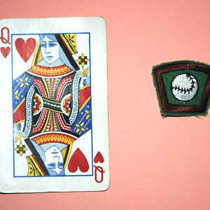 Pu&ograve; includere: Una carta da gioco che rappresenta la Regina di Cuori. La carta &egrave; rossa e bianca con un design nero e oro. La Regina &egrave; raffigurata con un cuore rosso in mano. Uno stemma verde e marrone con una palla da baseball bianca &egrave; sullo sfondo.