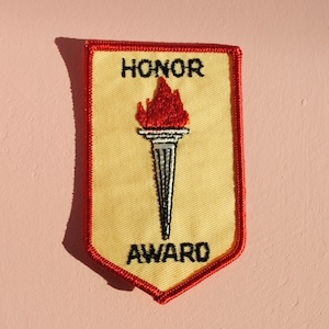 May include: A yellow patch with a red border and the words "HONOR AWARD" embroidered in black. A torch with a red flame is in the center. The patch is shield-shaped.
