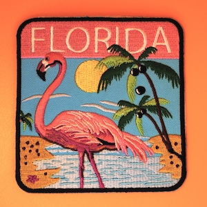 May include: Embroidered patch featuring a pink flamingo, palm trees, and a sun against a blue sky. The word "FLORIDA" is embroidered in white on a pink background. The patch has a black border.