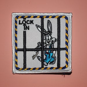 May include: A white embroidered patch with a blue and yellow border. The patch features a cartoon rabbit in a blue jumpsuit, holding a key, trying to escape from a jail cell. The text "LOCK IN" is embroidered in black on the patch.
