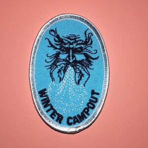 May include: A blue and white embroidered patch with a black and white illustration of a winter spirit blowing snow. The text "WINTER CAMPOUT" is written around the edge of the patch.