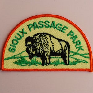 May include: A yellow and green embroidered patch with an orange border. The patch features a black silhouette of a bison and the text "Sioux Passage Park".