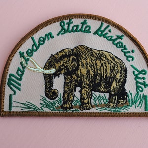May include: A brown and white embroidered patch with a green border. The patch features a brown mammoth with white tusks and the text "Mastodon State Historic Site".