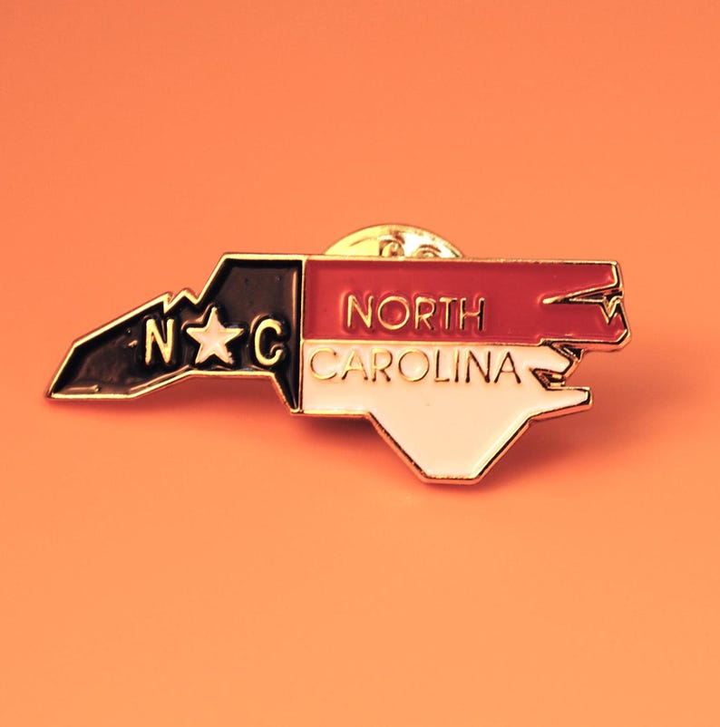 May include: A North Carolina state-shaped pin with a gold border. The pin features the state's outline, divided into sections of black, red, and white. The text "NORTH CAROLINA" is displayed on the red and white sections.
