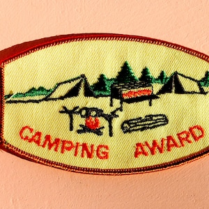 May include: A yellow and red embroidered patch with a camping scene. The patch features two tents, a campfire, and a log. The text "Camping Award" is embroidered in red on the patch.