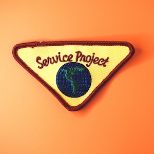 May include: A yellow triangular patch with a red border. The patch has a blue globe with green continents and the text "Service Project" in red.