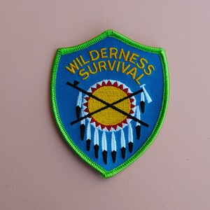 May include: A blue and green embroidered patch with the text "Wilderness Survival" and a Native American headdress design.