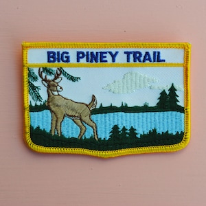 May include: A yellow and blue embroidered patch with a white background. The patch features a deer standing in a forest by a lake. The text "BIG PINEY TRAIL" is written in blue on the top of the patch.