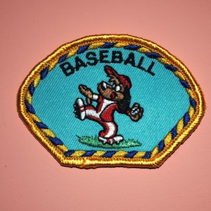 May include: A blue and yellow embroidered patch with a cartoon character playing baseball. The patch says "BASEBALL" in black letters.