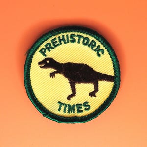 May include: Embroidered patch with a dinosaur silhouette inside a yellow circle, surrounded by the words "PREHISTORIC TIMES" in green. The patch has a green border and is set against an orange background.