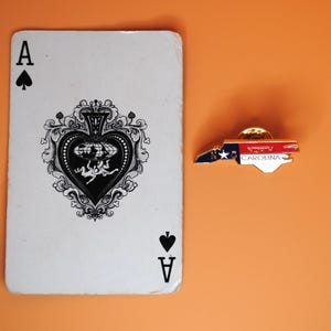 May include: An Ace of Spades playing card next to a North Carolina state-shaped pin. The pin features the state flag design with the text "NORTH CAROLINA". The background is a solid orange colour.
