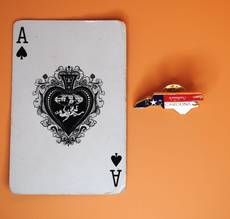 May include: An Ace of Spades playing card next to a North Carolina state-shaped pin. The pin features the state flag design with the text "NORTH CAROLINA". The background is a solid orange colour.