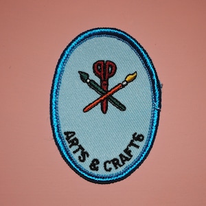May include: A blue oval-shaped patch with a white background and the text "Arts & Crafts" in black embroidery. The center of the patch features a red, green, and yellow embroidered design of a pair of scissors, a paintbrush, and a pencil.