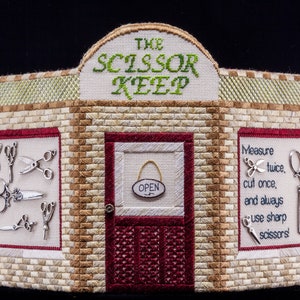 May include: A needlepoint wall hanging depicting a storefront with the words "The Scissor Keep" above the door. The storefront has a brick facade with two windows and a red door with an "Open" sign. The windows display various scissors and a quote about measuring twice and cutting once.