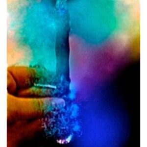 May include: A hand holds a glass object, with a cloud of blue, green, and purple smoke emanating from it. The background is dark, with the smoke illuminated by a light source.