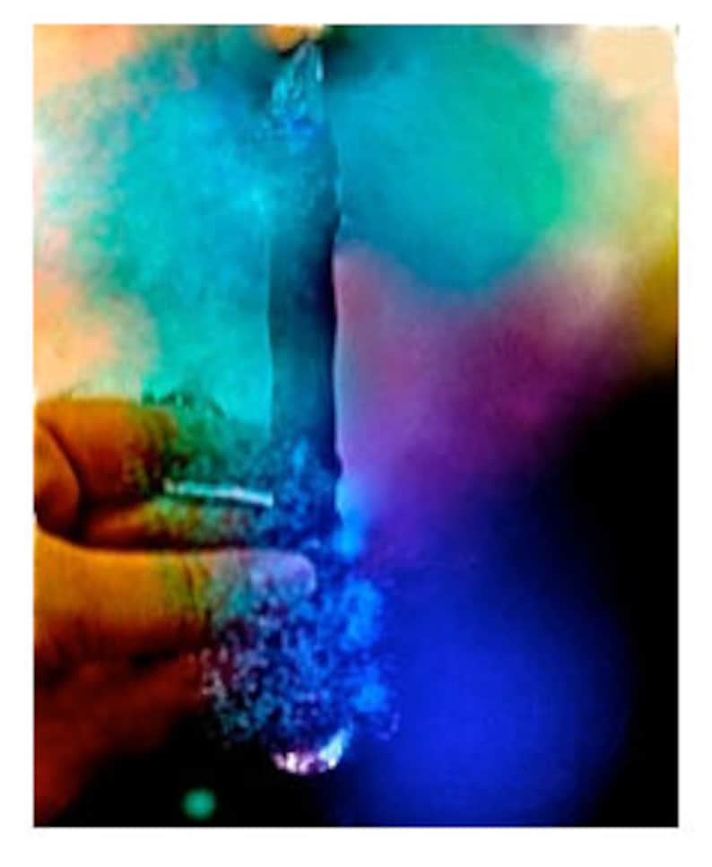 May include: A hand holds a glass object, with a cloud of blue, green, and purple smoke emanating from it. The background is dark, with the smoke illuminated by a light source.
