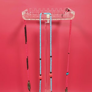 May include: Three beaded necklaces displayed on a clear acrylic stand. The necklaces are made of black, pink, and turquoise beads. The turquoise necklace has two larger pink beads and a brown pendant.