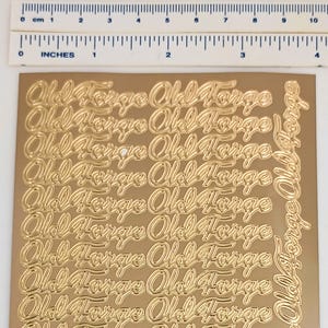May include: A sheet of gold-colored stickers with the words "Old Forge" in a cursive font. The stickers are arranged in rows and columns. The ruler at the top measures up to 4 inches.
