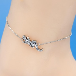 May include: A silver-colored anklet featuring a detailed lizard charm. The lizard is intricately designed with textured scales and a curved tail. The anklet chain is delicate and attached to the lizard charm.