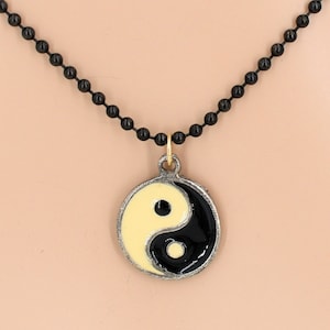 May include: A black beaded chain necklace with a silver pendant featuring a yin yang symbol. The yin yang symbol is painted in black and cream.