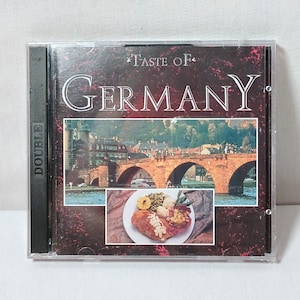 May include: A CD case with a red background and the text "Taste of Germany" in white. The front cover features a photo of a bridge over a river with a cityscape in the background. The bottom of the cover features a photo of a plate of food with a chicken breast, potatoes, and green beans.
