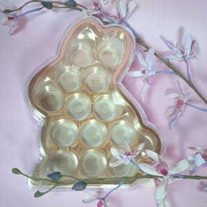 May include: A clear plastic bunny-shaped container with multiple circular compartments, possibly for chocolates or sweets. The container has a gold-coloured interior and a pinkish-gold rim. The background is a soft pink, with delicate white and purple orchid blossoms.