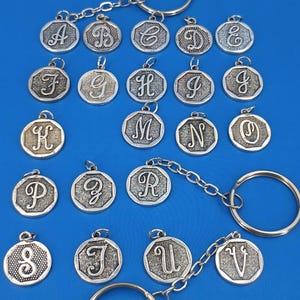 May include: Silver-colored metal keychains with various engraved letters of the alphabet. Each charm is an octagon shape with a loop for attaching to a keyring. The keychains are displayed on a blue background.